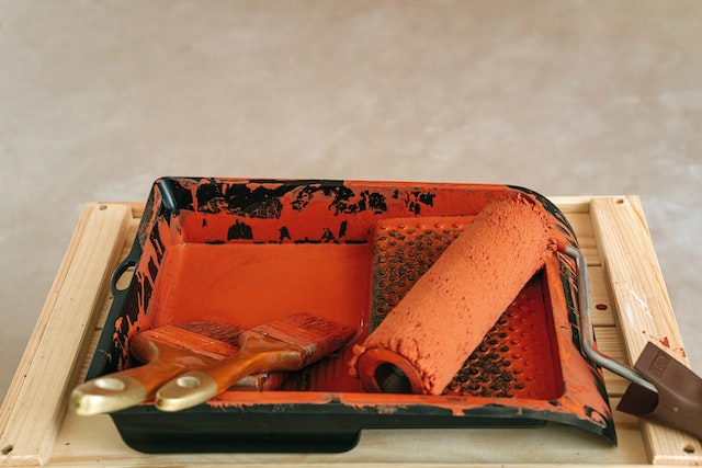 two paint brushes and a roller in a tray with orange paint